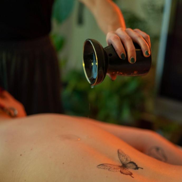 Joy giet warme massageolie op de rug van een klant met vlindertatoeage
