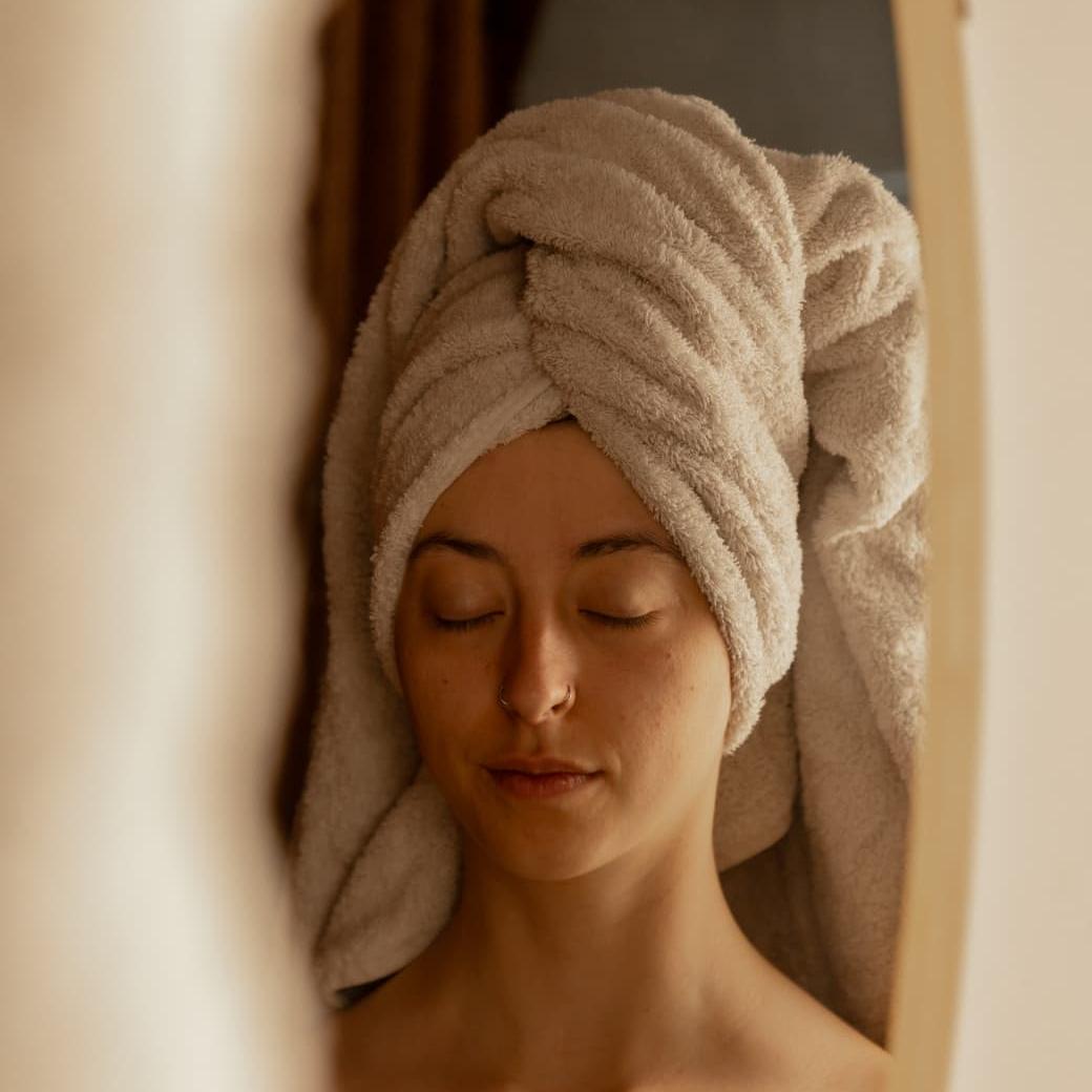 Vrouw met handdoektulband kijkt ontspannen in de spiegel na wellnessbehandeling